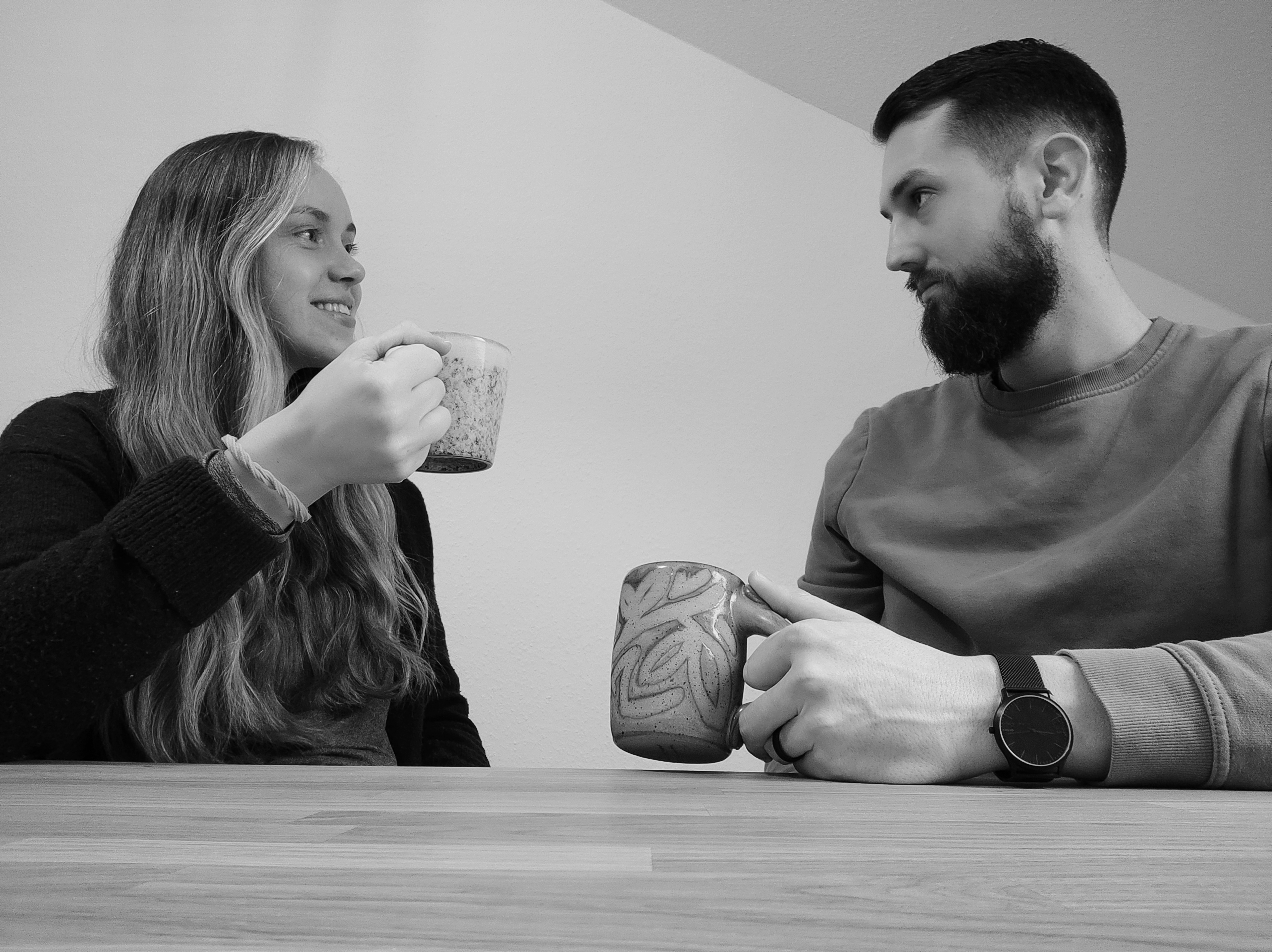 A woman with long, blonde hair and a man with short, dark hair are sitting at a table with their coffee cups. They are involved in a conversation.