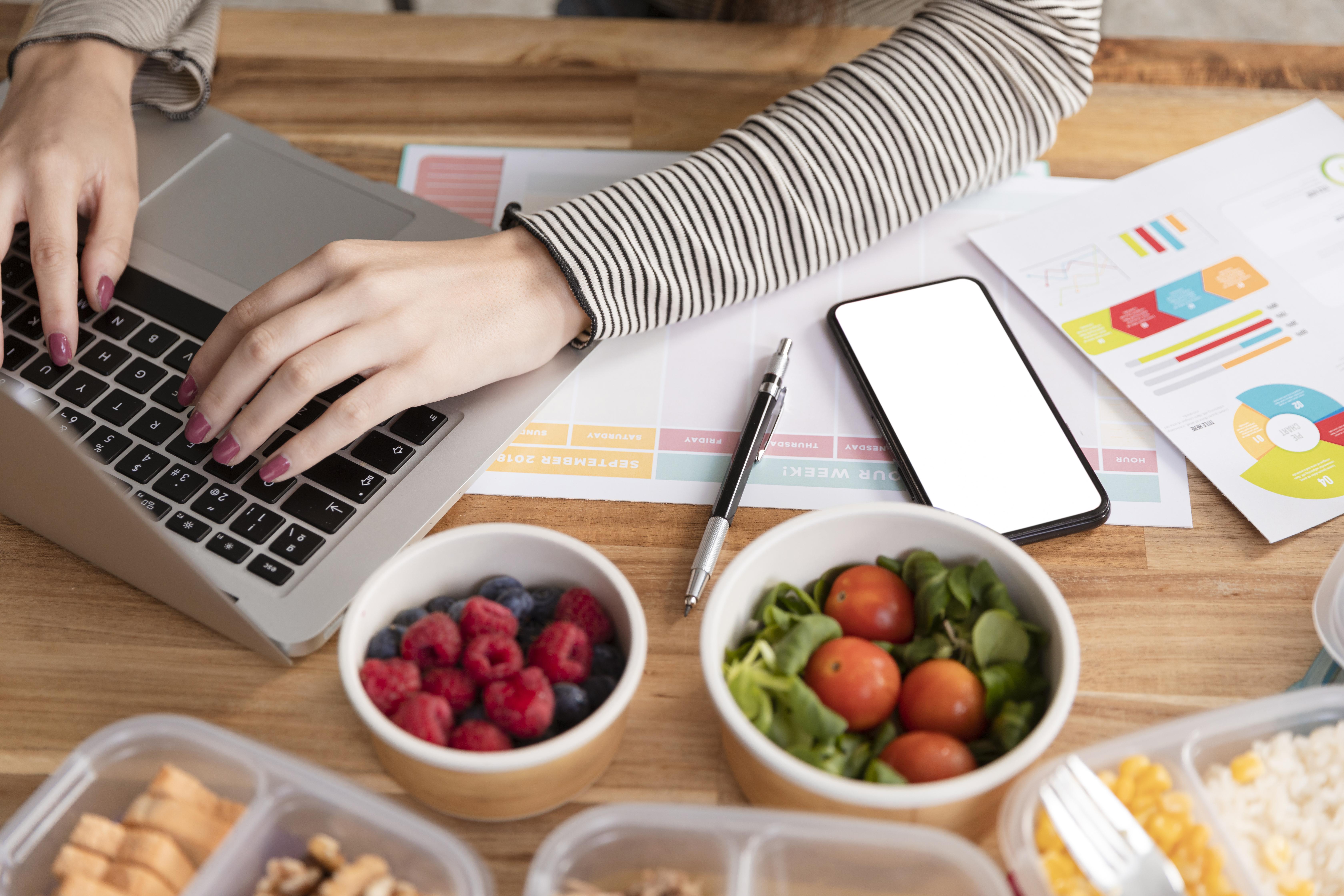 Arme einer am Laptop arbeitenden Person mit Essen und Unterlagen auf dem Tisch